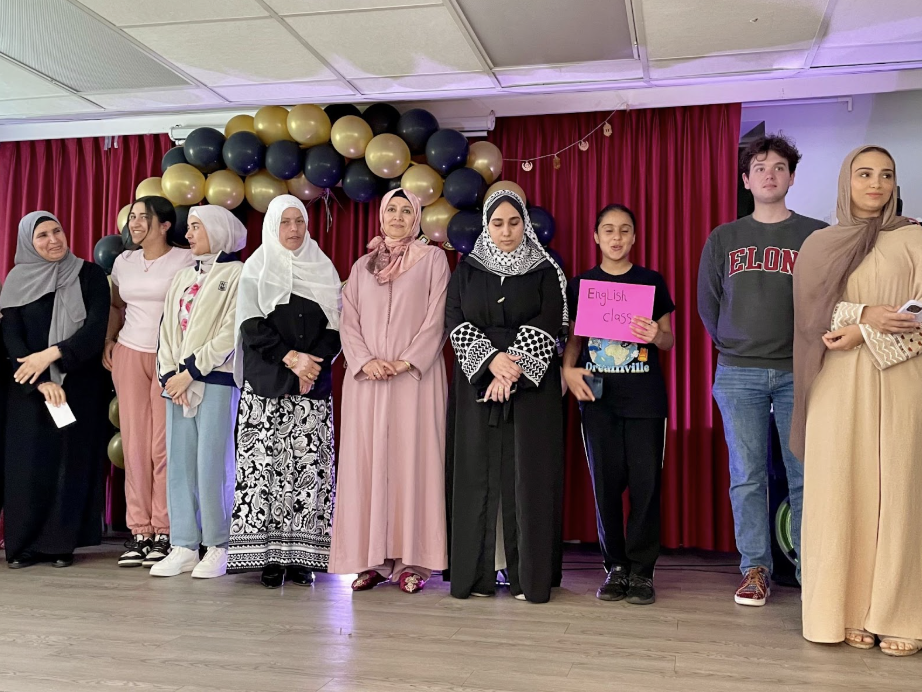 A diverse group of women and students stand on stage in front of a red curtain and balloon arch, with one holding a sign that reads “English class.